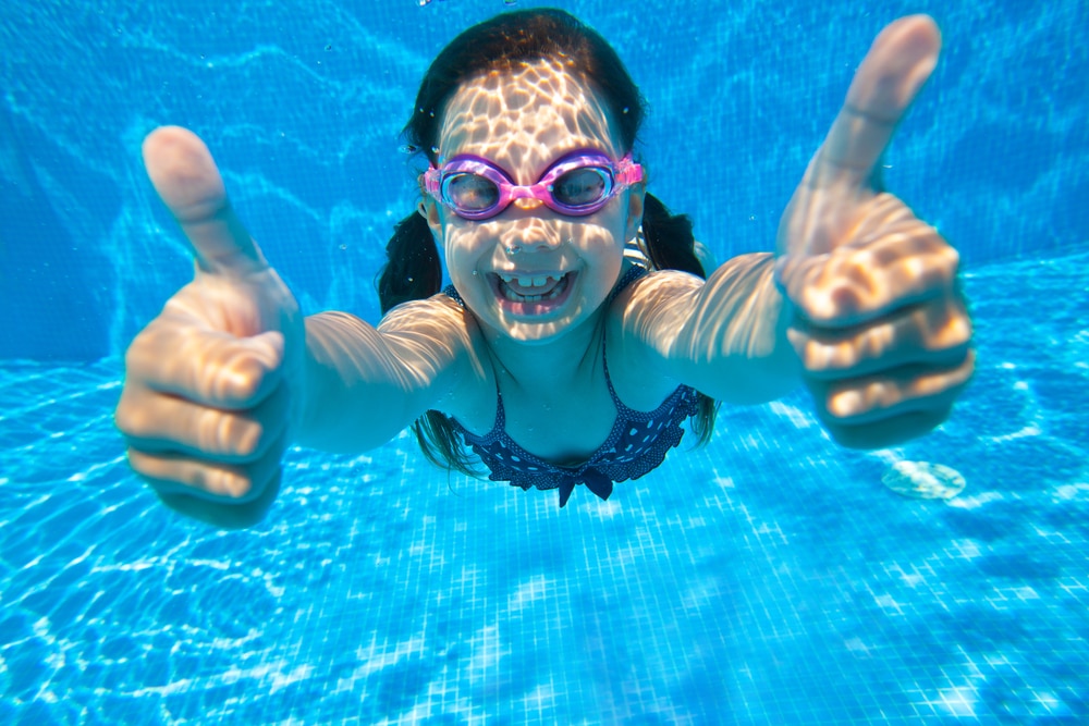 A young girl wearing pink goggles gives two thumbs up while smiling underwater in a clear blue swimming pool, heated by energy-efficient heat pumps. The sunlight patterns dance on her skin. | AES - Pool Heating & Energy Efficient Products