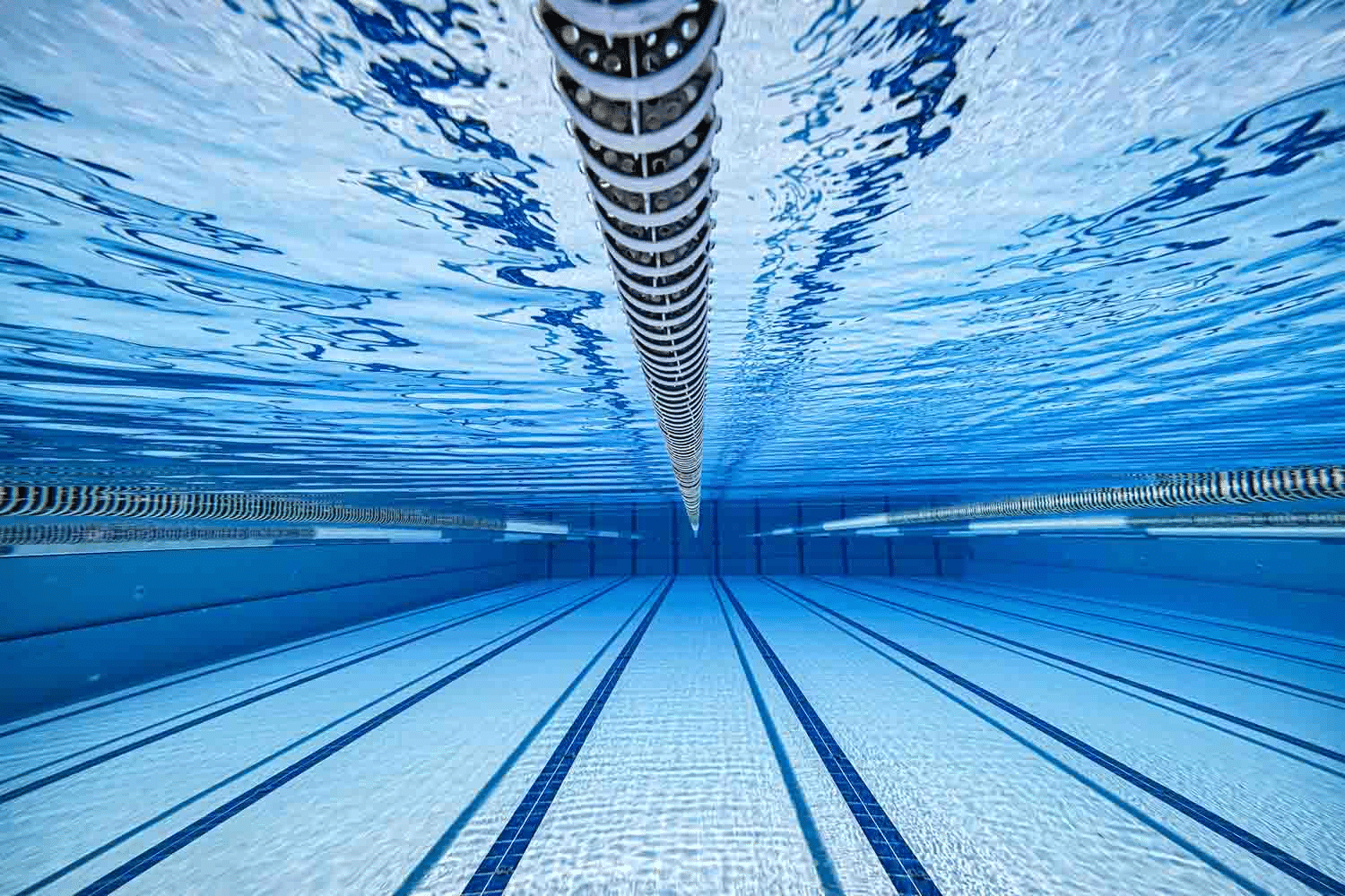 Underwater view of a clear, blue swimming pool showing lane markers and the ripple of water on the surface, with lane ropes extending toward the horizon. | AES - Pool Heating & Energy Efficient Products