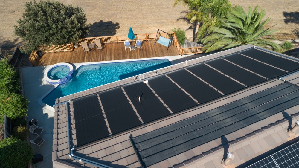 Aerial view of a home with solar panels on the roof, featuring an adjacent swimming pool with lounge chairs and a palm tree. | AES - Pool Heating & Energy Efficient Products
