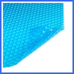 Close-up image of a blue pool cover with a textured, bubble-like surface. One corner of the cover is slightly lifted, revealing the flexible material. The cover appears to be made of plastic and designed to float on top of a swimming pool. | AES - Pool Heating & Energy Efficient Products